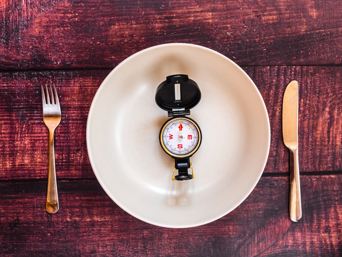 An empty plate with a compass in the center, flanked by a fork and knife on a wooden table, symbolizing direction and decision-making in intermittent fasting and weight loss.
