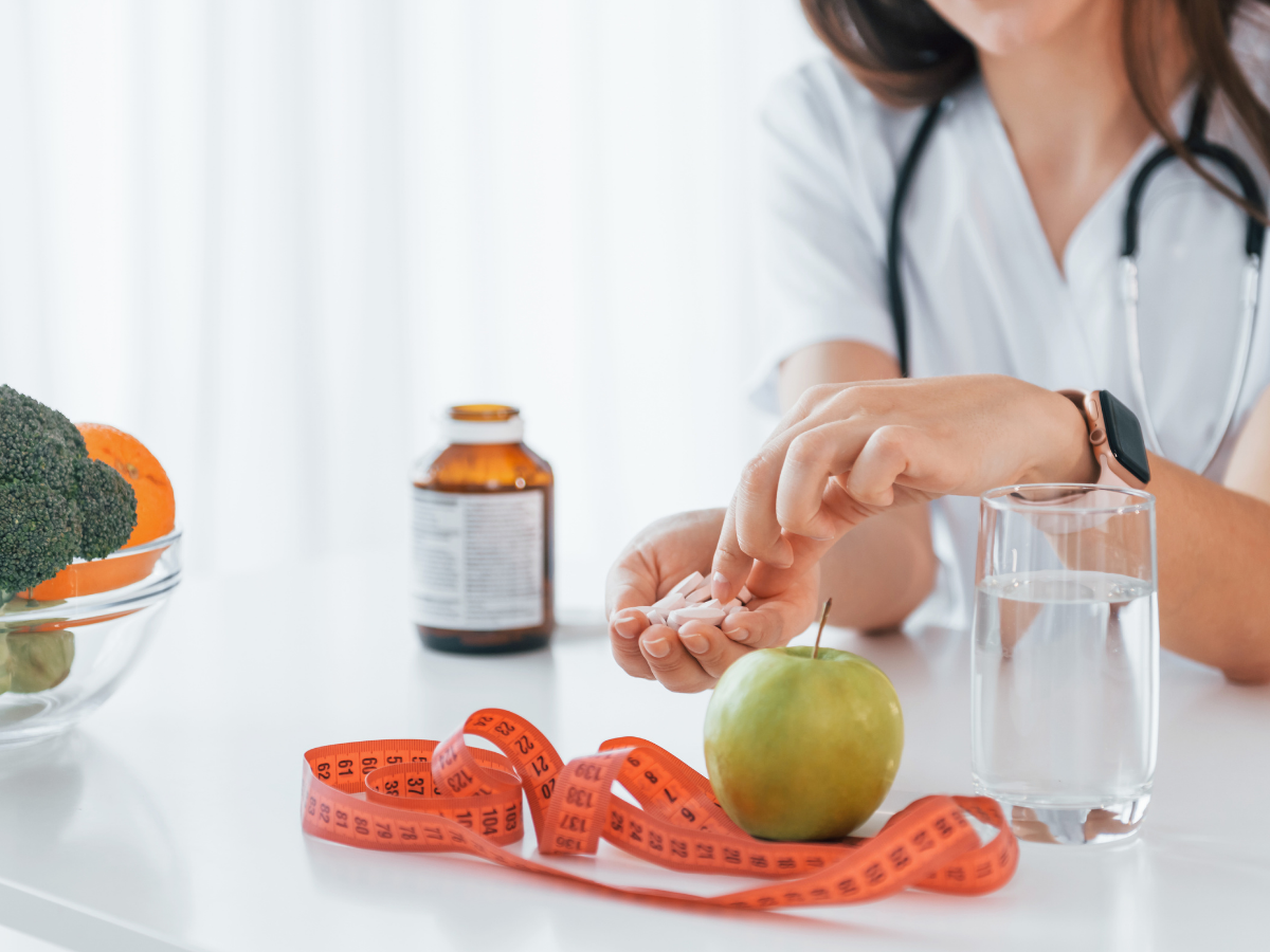 A healthcare professional holding white tablets beside a green apple, glass of water, and orange measuring tape on a white table, suggesting weight management through medication and nutrition.