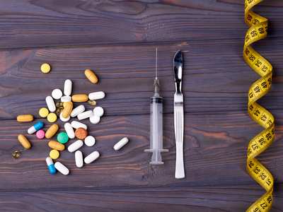 Various weight loss tools on a wooden surface, including pills, a syringe, a surgical scalpel, and a yellow measuring tape. Visually representing different approaches to weight loss treatments like Saxenda and Ozempic.