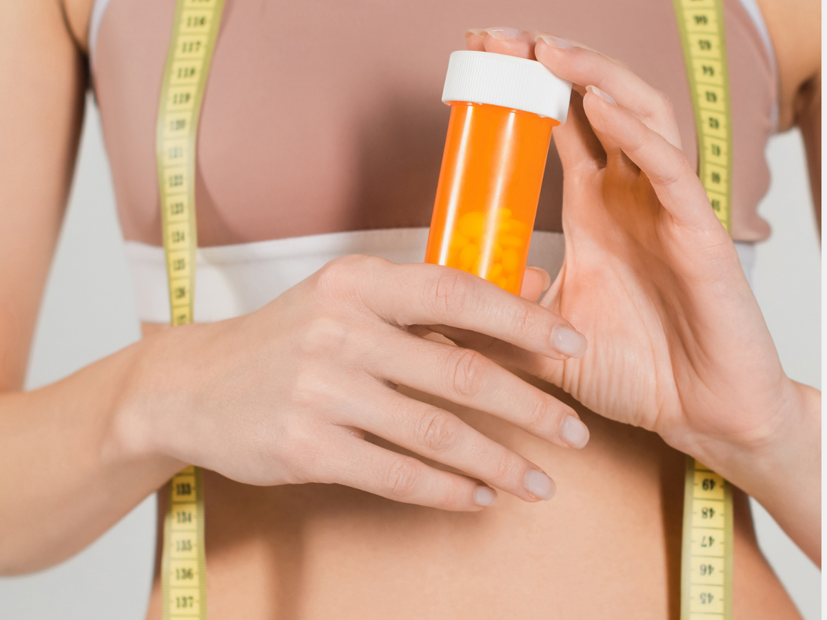Close-up of a fit woman holding a pill bottle with a measuring tape around her waist, symbolizing the comparison of medical weight loss treatments like Semaglutide and Tirzepatide.